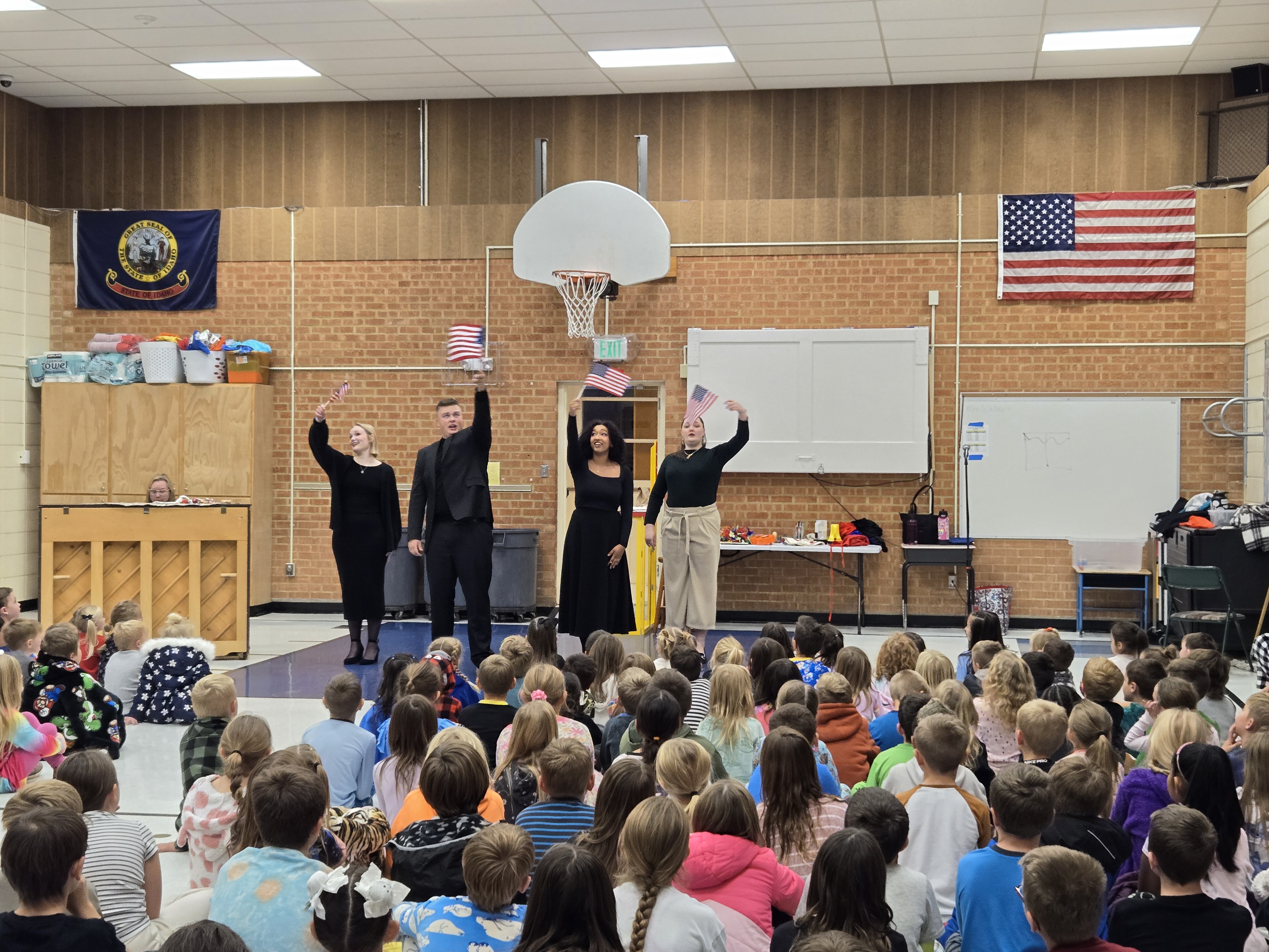 Four performers wave American flags on a school gym stage, facing an audience of seated children.