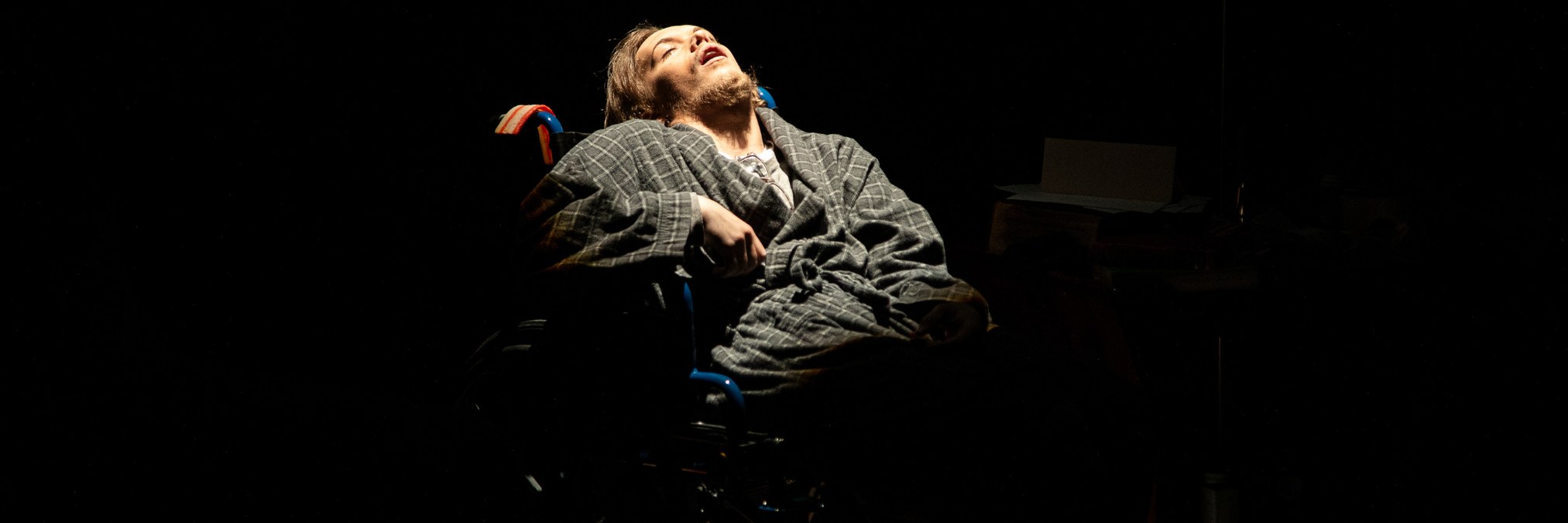 Black background, with man in bathrobe leaning his head back against the wheelchair he is sitting in