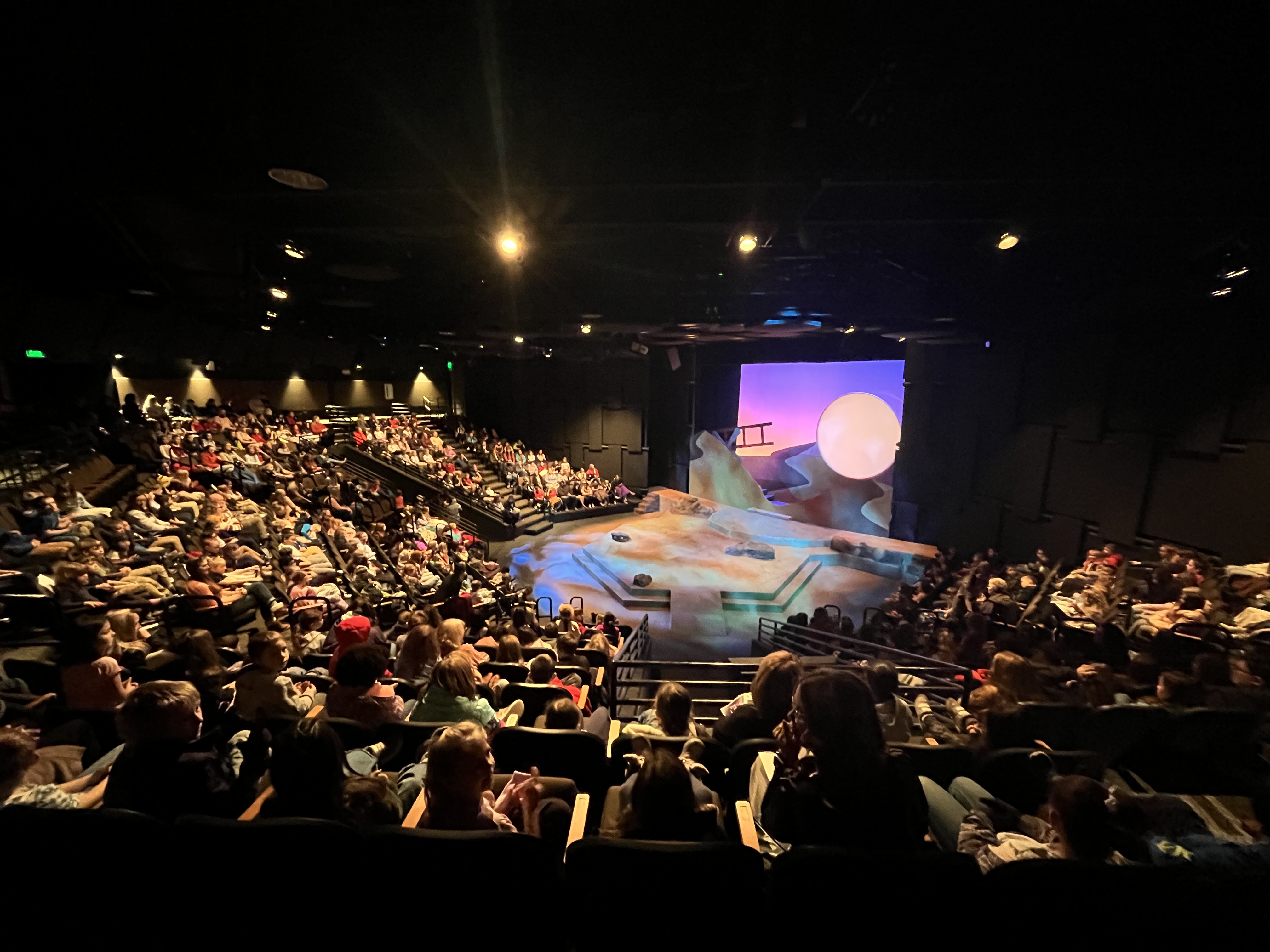 Hundreds of elementary school students fill the auditorium seats overlooking a stage with pastel colored lighting and a large full moon.