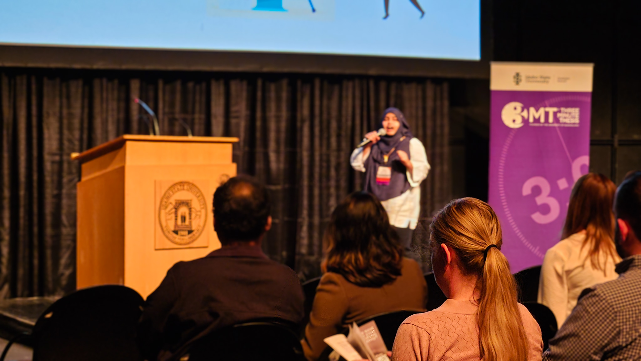 Audience watching a graduate student present at the 3MT event