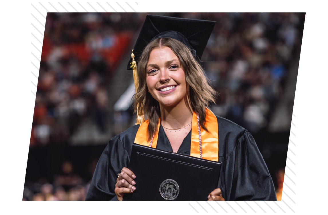 Graduate wearing a black cap and gown smiles while holding a diploma cover during a commencement ceremony, with a blurred audience in the background.