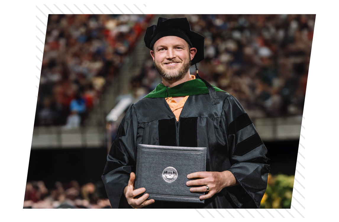 Graduate wearing academic regalia and a black doctoral cap holds a diploma folder while standing in a large indoor arena with a blurred audience in the background.