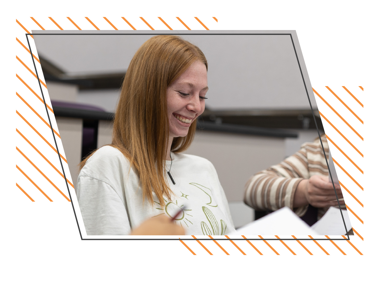 Smiling college student seated in a classroom, looking down at paperwork while participating in a small group discussion, representing an engaged learning environment.