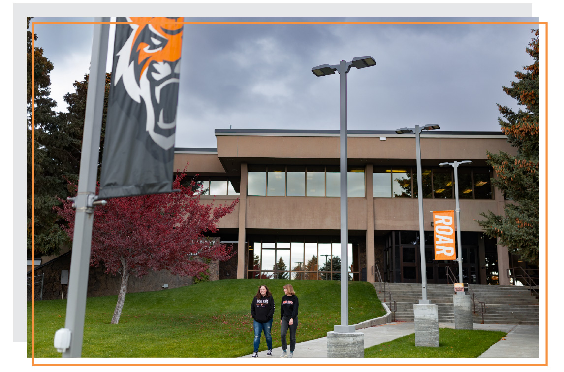 Two students walking on a sidewalk in front of an ISU campus building, with ISU Bengal banners and fall trees visible nearby.