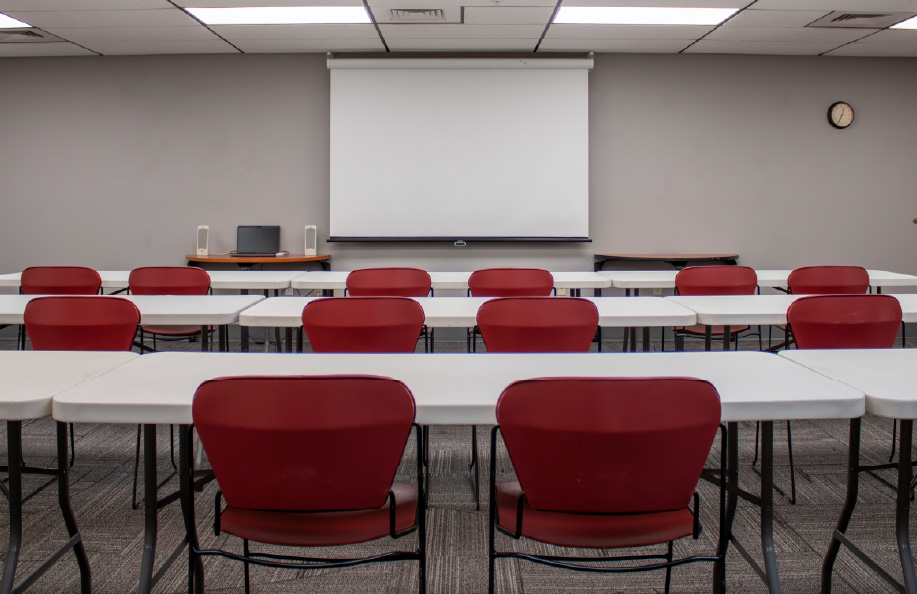Classroom with rows of white tables and red chairs facing a ceiling-mounted projector and pull-down screen, with a laptop and speakers on a front table and a wall clock visible.