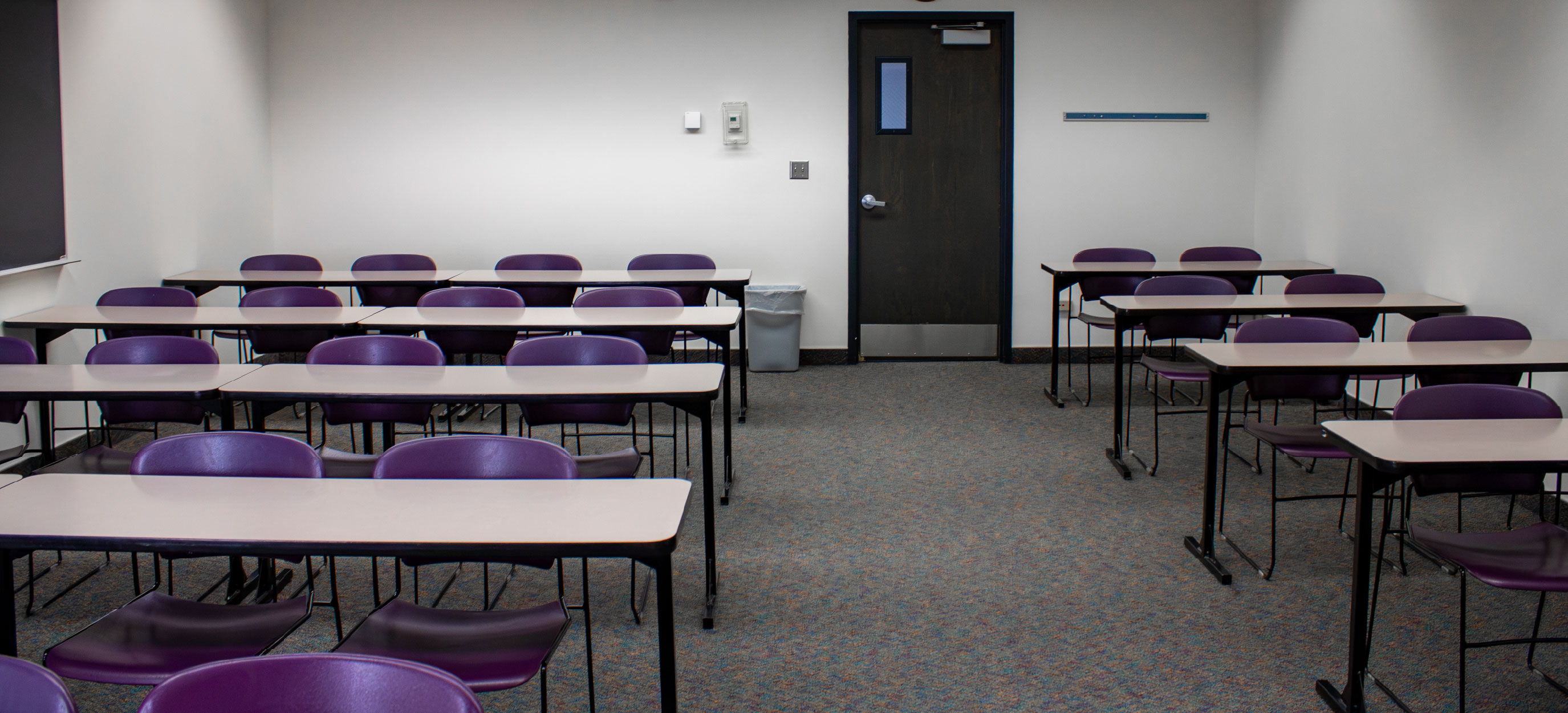 CHE classroom with rows of tables and purple chairs arranged around a central aisle, with a doorway at the back of the room.