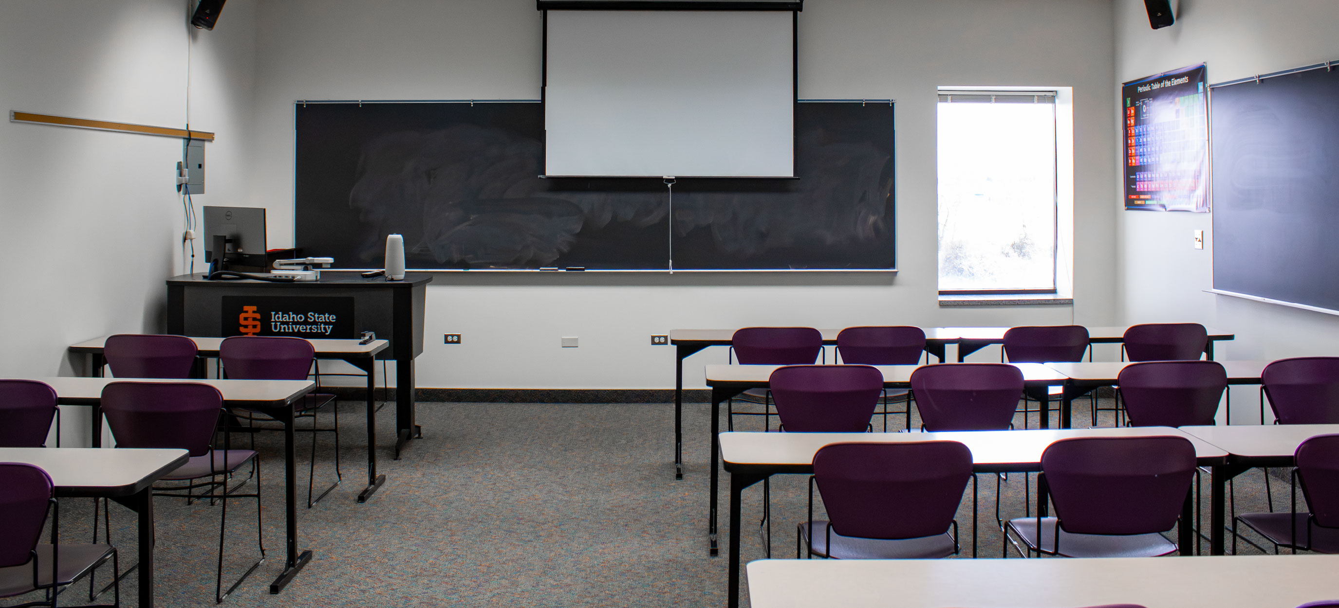 CHE classroom with rows of tables and purple chairs facing a teaching station, large chalkboards, and a pull-down projection screen.