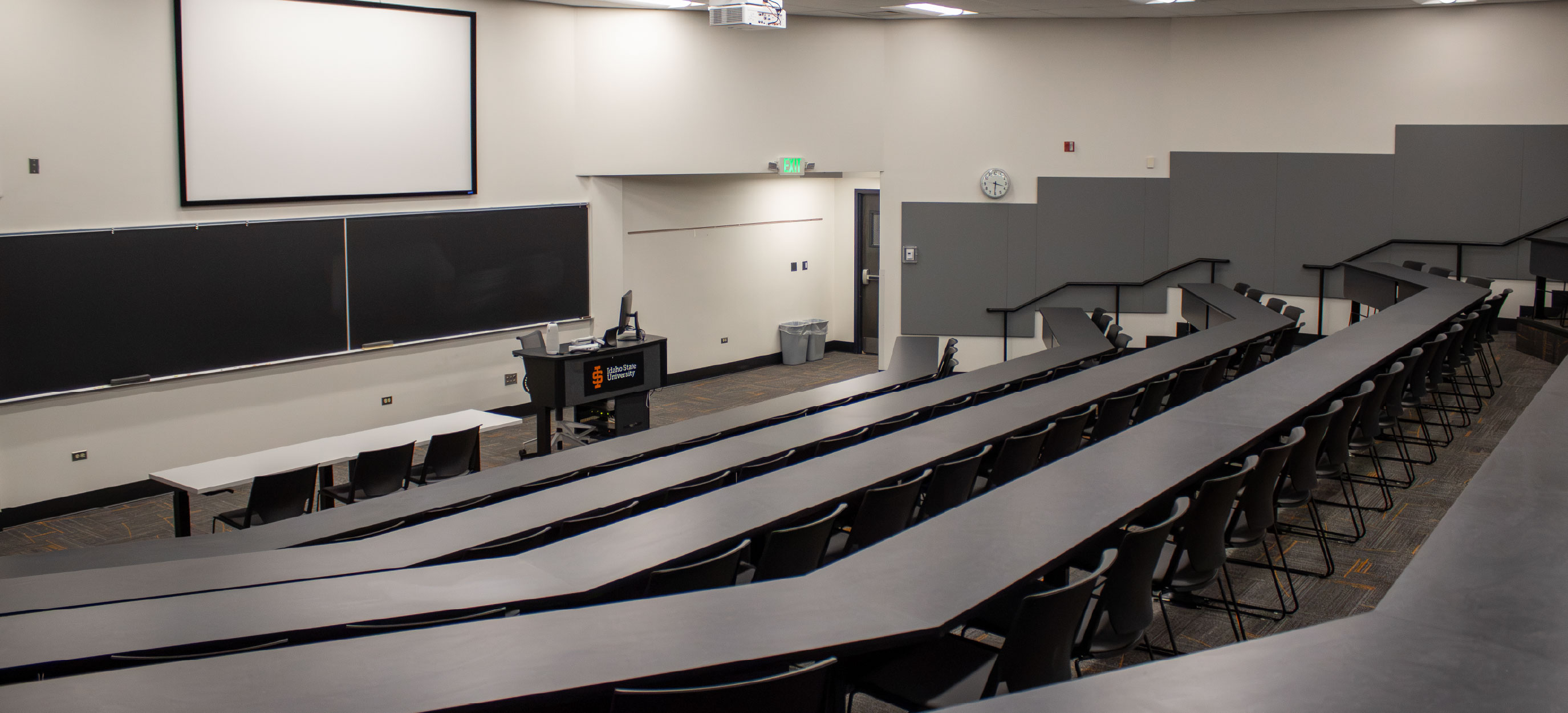 Wide view of the CHE Large Tiered Classroom at ISU Idaho Falls, showing curved rows of fixed tables and chairs facing blackboards, a projection screen, and an instructor station.