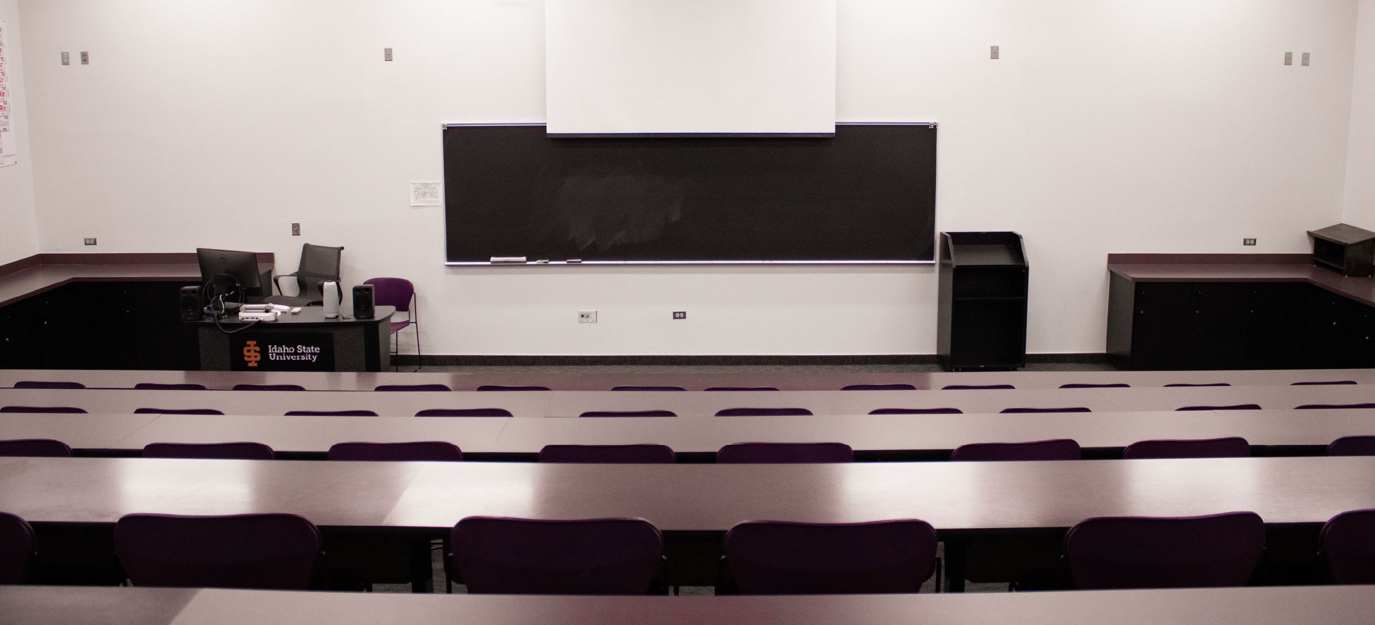 Front view of the CHE Tiered Classroom at ISU Idaho Falls, showing tiered tables and purple chairs facing a large blackboard, projection screen, and instructor station.
