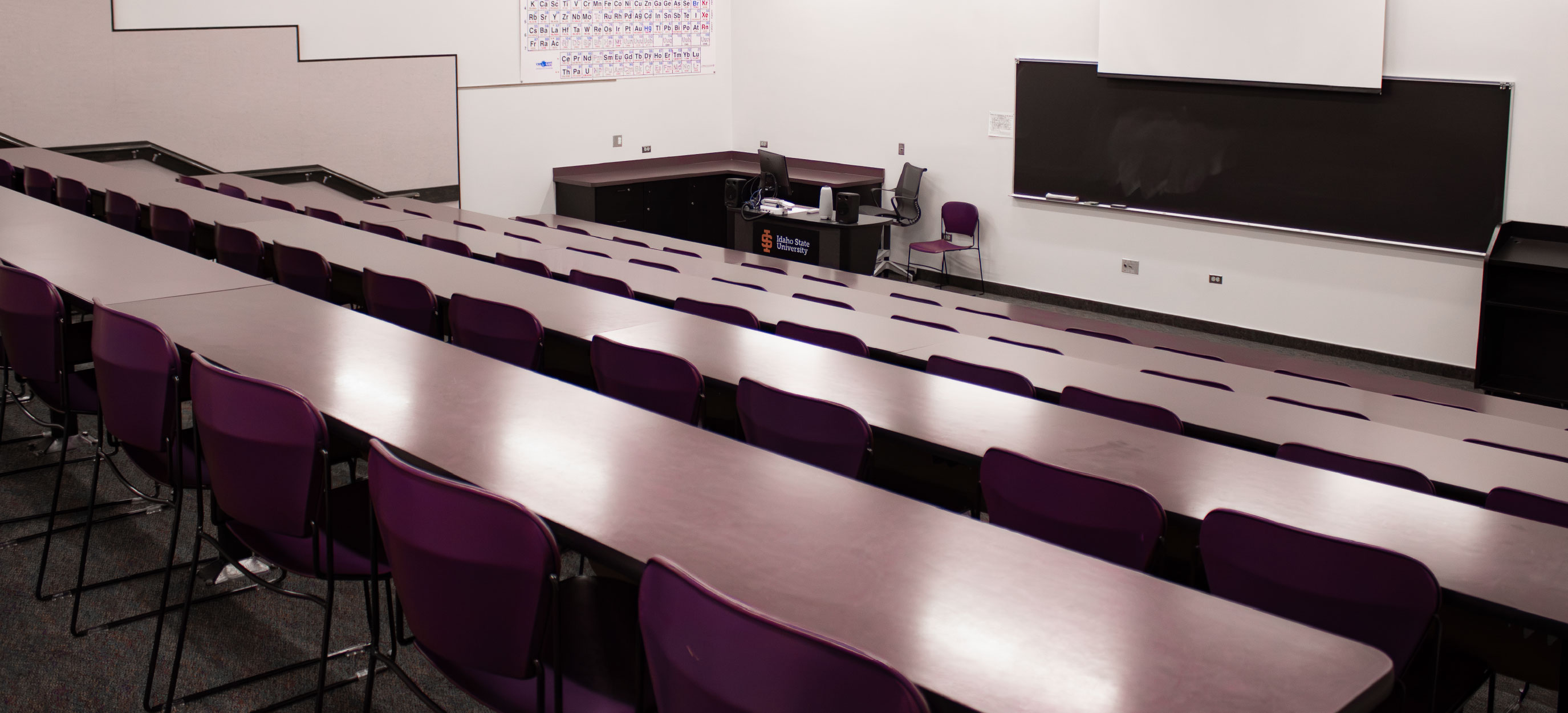 Wide view of the CHE Tiered Classroom at ISU Idaho Falls, showing rows of fixed tables and purple chairs facing the instructor station, blackboard, and projection screen.