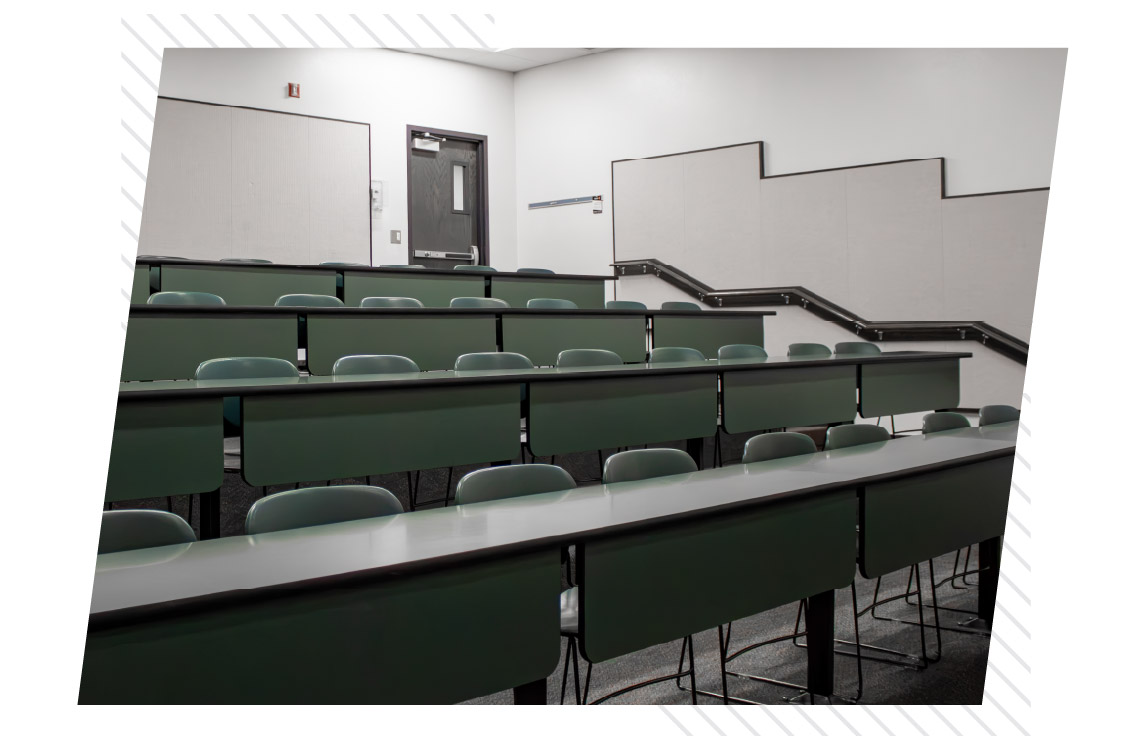 Small tiered CHE classroom with fixed rows of green seating and long desks arranged on ascending levels, with stairs and handrails along the side.