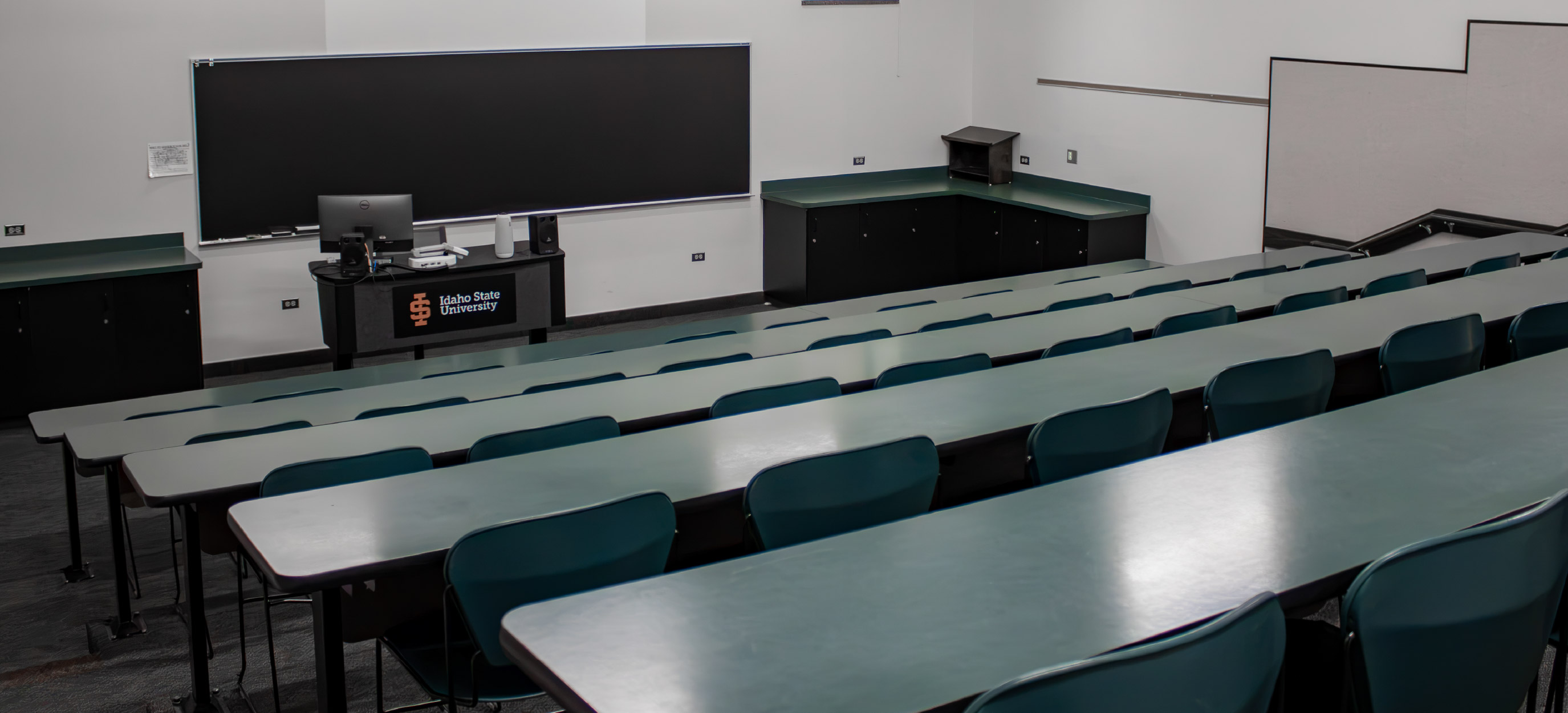 Small CHE tiered classroom with rows of fixed desks and green chairs facing a teaching station and large chalkboard at the front of the room.