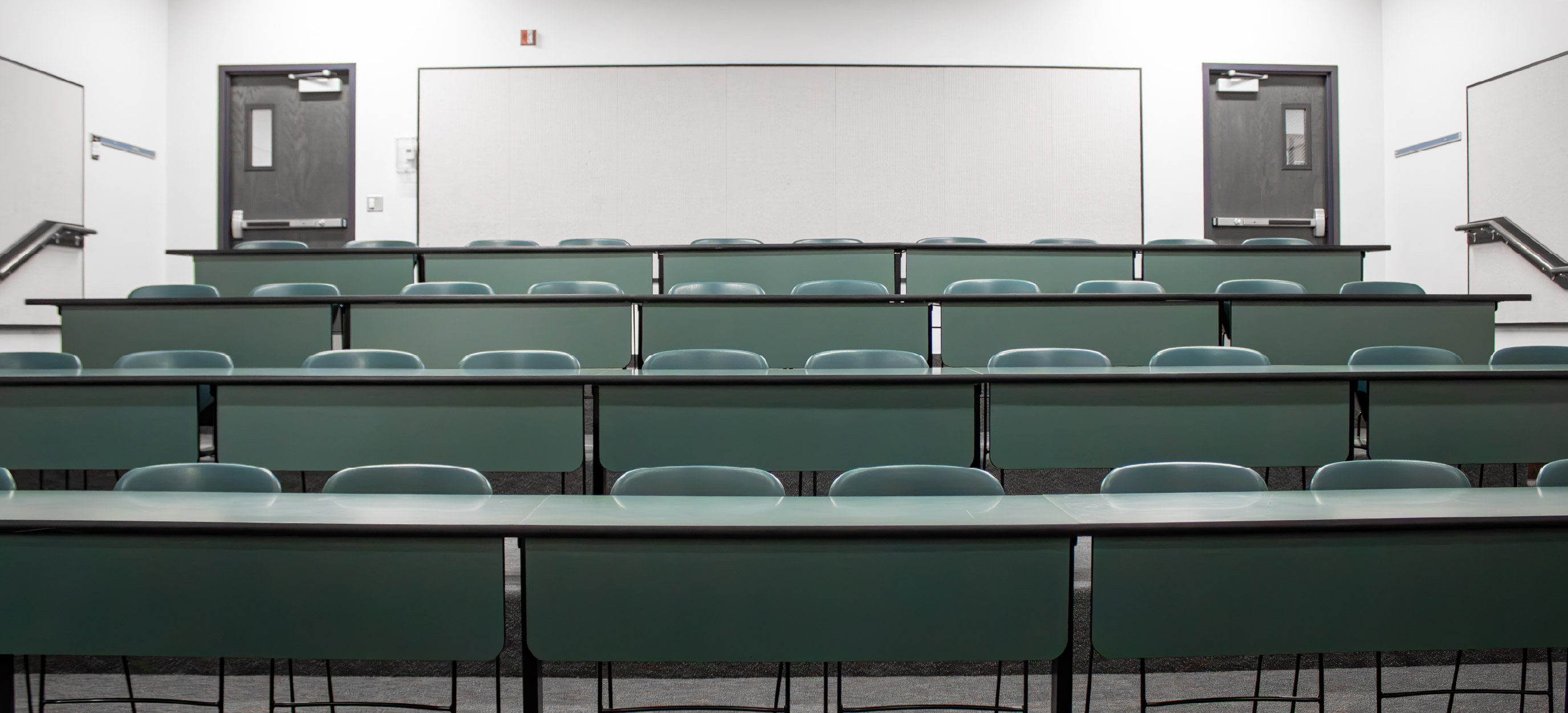 Small CHE tiered classroom viewed from the front, showing multiple rows of fixed green desks and chairs arranged on ascending levels with doors at the back.