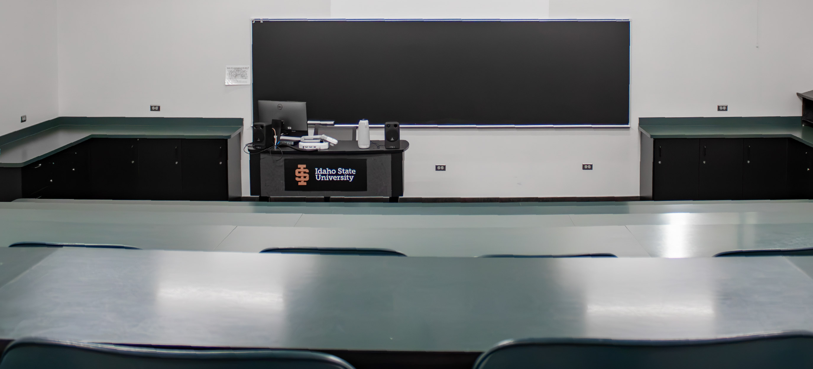 View from the tiered seating toward the front of the small CHE classroom, showing long desks, a teaching station with technology, and a large chalkboard.