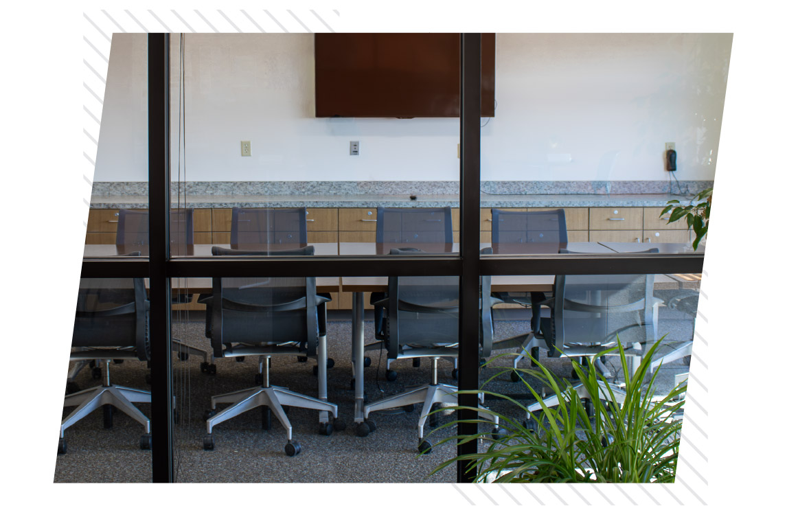 Conference room viewed through glass walls, with a long table, black office chairs, wall-mounted monitor, and plants.