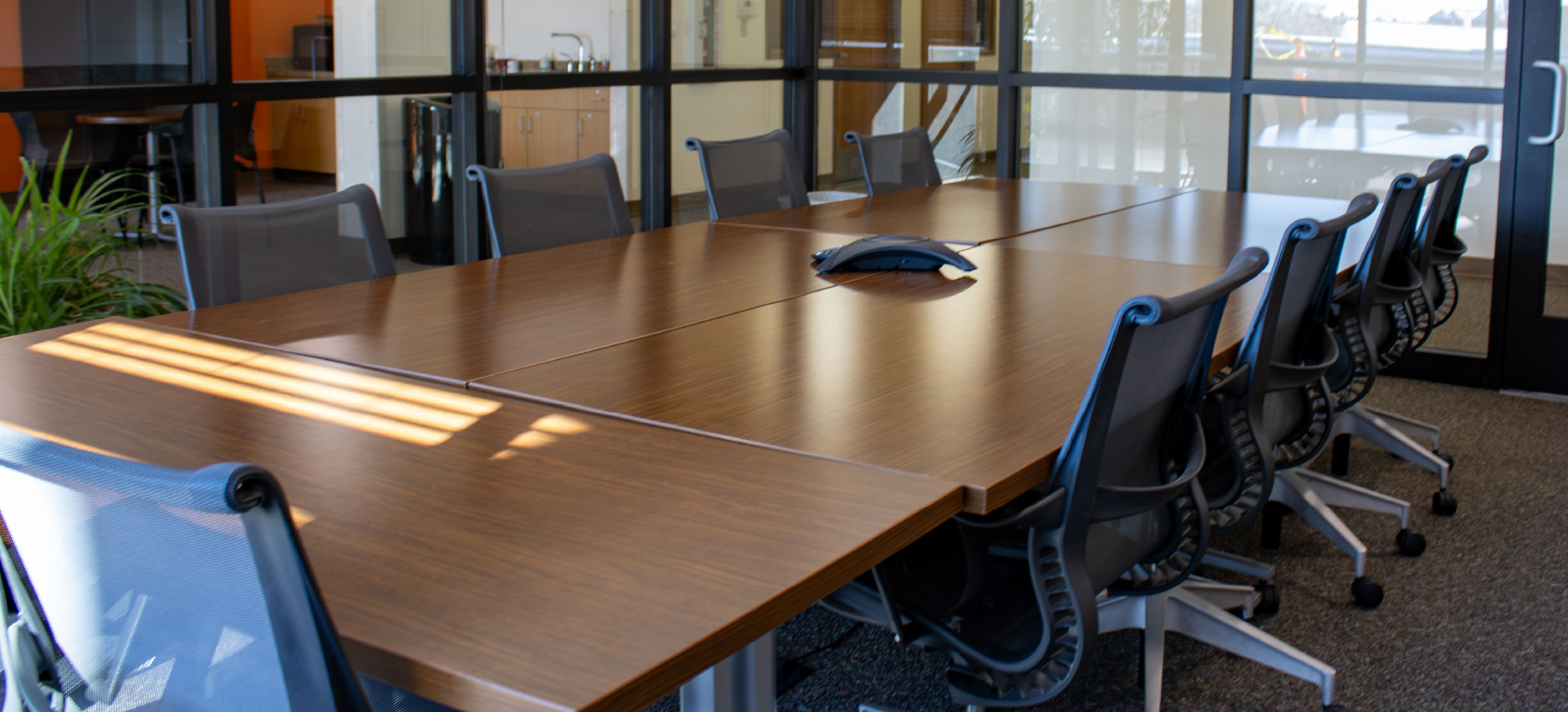 Conference room featuring a large wooden table, black rolling chairs, a conference phone, and glass walls surrounding the space.