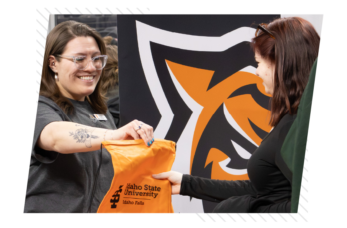 Staff member handing an Idaho State University Idaho Falls drawstring bag to a visitor in front of a Bengal-themed backdrop at a campus event.