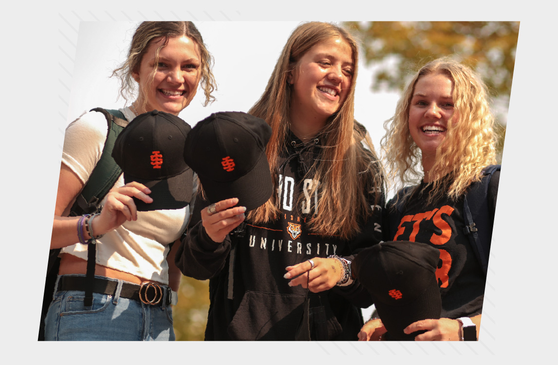Three female college students stand outdoors on campus, smiling and holding black baseball caps with an orange ISU logo. They wear casual clothing and backpacks, with fall trees softly blurred in the background, creating a warm and welcoming university atmosphere.