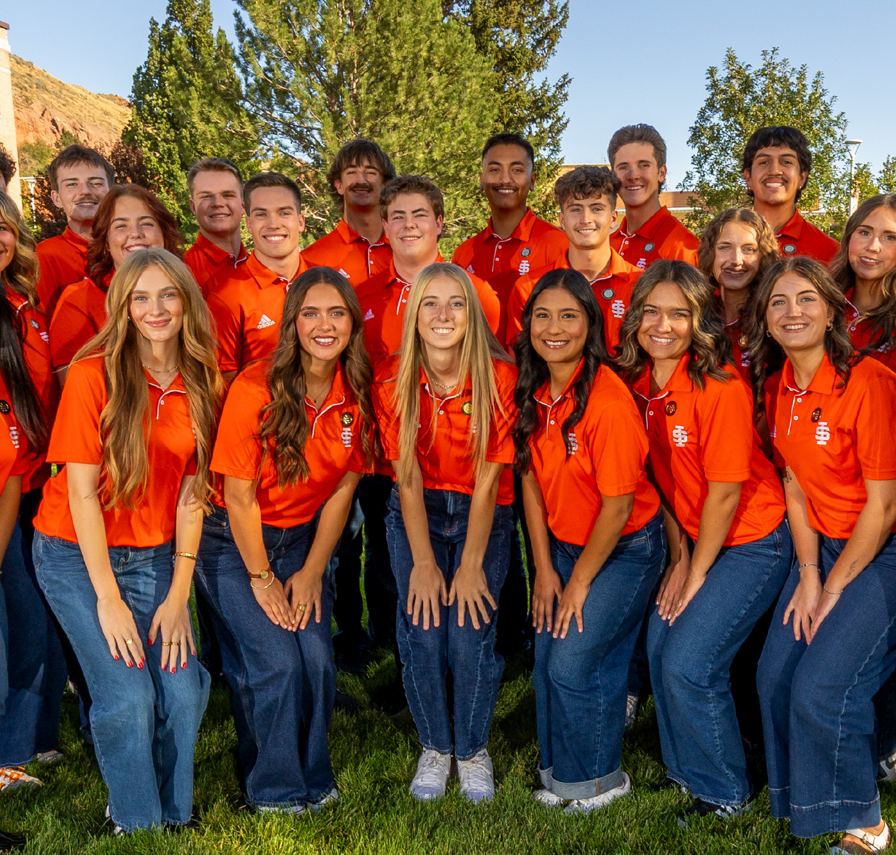 A large group of college students stand together outdoors on campus, smiling for a group photo. They wear matching orange ISU polo shirts and jeans, with trees and campus buildings in the background, creating a strong sense of school pride and student involvement.