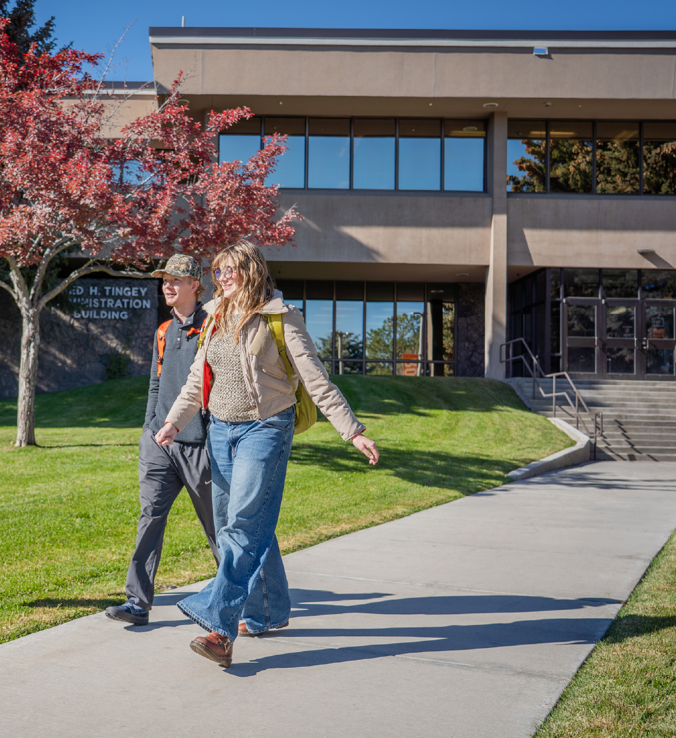 Two college students walk together on a sunny campus sidewalk in front of the Administration Building. A red-leaved tree and green lawn frame the scene as they carry backpacks and talk, creating a welcoming and active campus atmosphere.