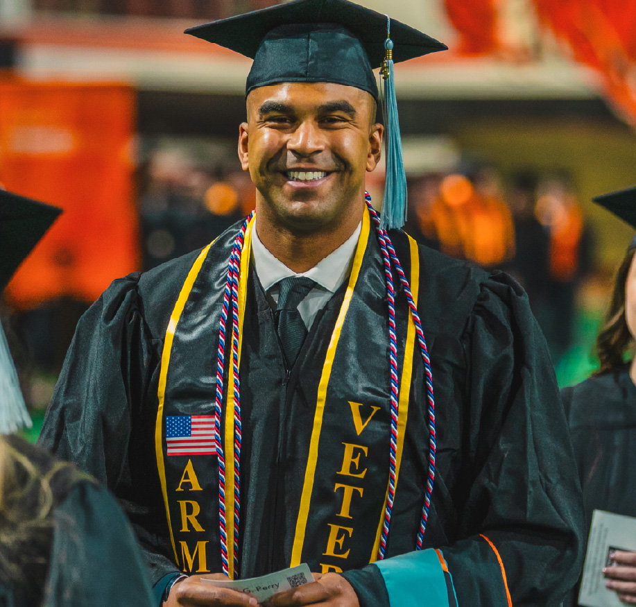 A smiling graduate stands in cap and gown during a commencement ceremony. The graduate wears honor cords and a stole that reads “Army Veteran,” holding a program while celebrating the achievement.