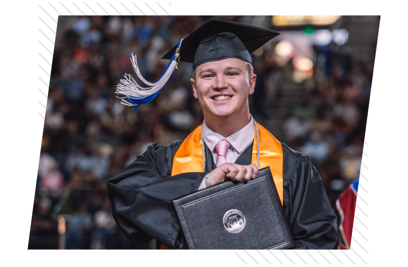 Graduate wearing a black cap and gown smiles while holding a diploma cover during a commencement ceremony, with a crowd visible in the background.