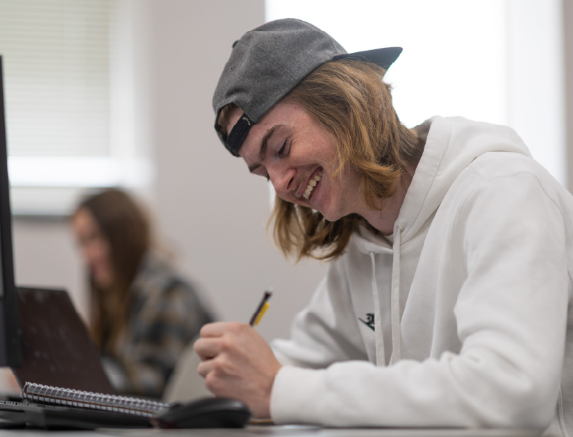 Student wearing a hoodie and backward baseball cap smiles while writing in a notebook at a desk. Another student works on a laptop in the background of a classroom.