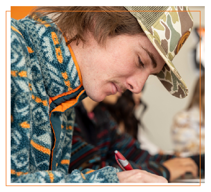 Student wearing a hat and patterned jacket writes on paperwork in a classroom, representing the process of applying to Idaho State University.