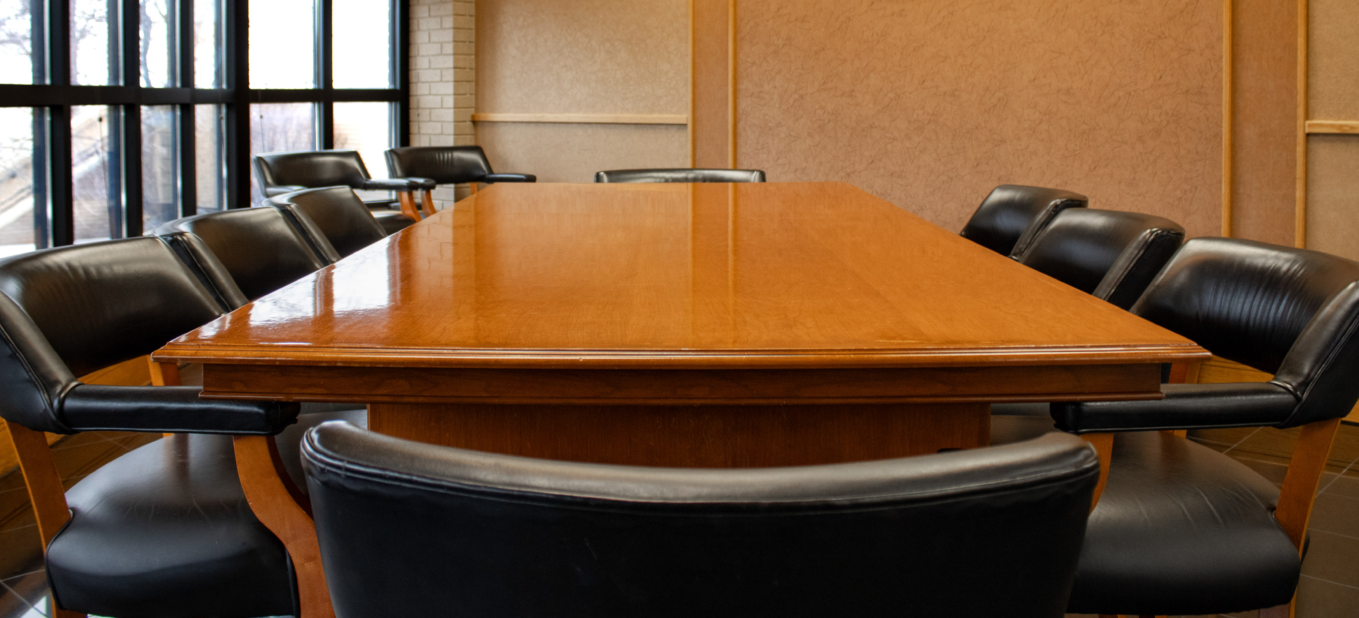 Conference room with a polished wooden table surrounded by black leather chairs, with large windows along one side.