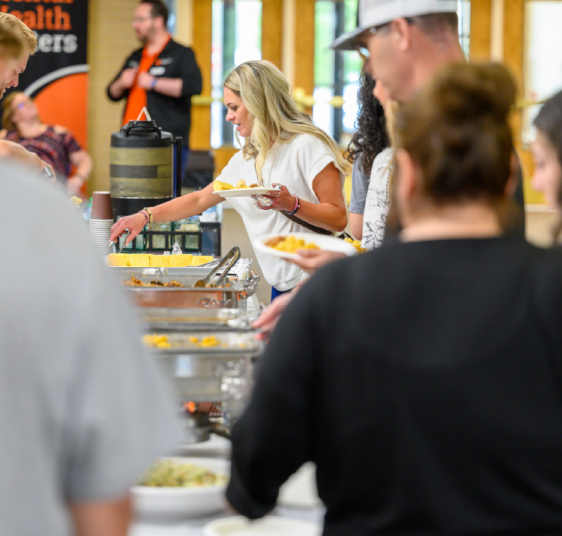 People serve themselves food from a buffet line at a campus event, with a woman reaching for food while holding a plate and others gathered around the serving tables.