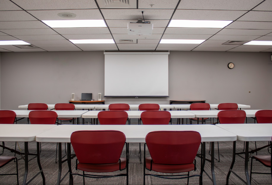 Classroom with rows of white tables and red chairs facing a ceiling-mounted projector and pull-down screen, with a laptop and speakers on a front table and a wall clock visible.