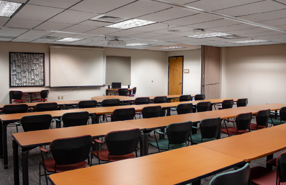Classroom with rows of long wooden tables and black chairs facing a projection screen and instructor desk at the front, with wall-mounted artwork and a clock visible.