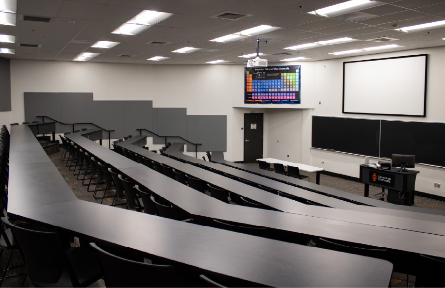Large tiered lecture hall with curved rows of fixed desks and chairs arranged stadium-style, facing a projection screen, chalkboards, and instructor podium at the front of the room.