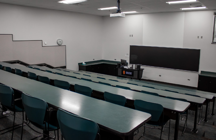 Tiered lecture classroom with rows of long tables and green chairs arranged stadium-style, facing a chalkboard, projection screen, and instructor podium at the front.