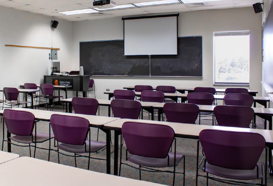 Classroom with rows of tables and purple chairs facing a large projection screen and blackboards at the front of the room. An instructor station, ceiling-mounted projector, speakers, and large windows are also visible.