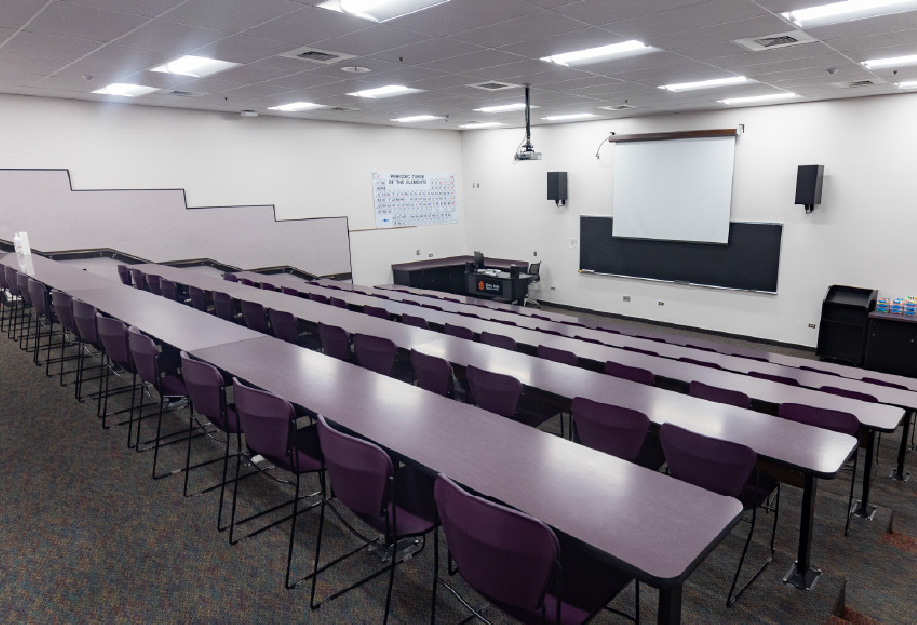 A medium-sized tiered classroom with rows of long tables and purple chairs facing the front of the room. The classroom includes a projector and screen, chalkboard, instructor station, wall-mounted speakers, and overhead lighting.