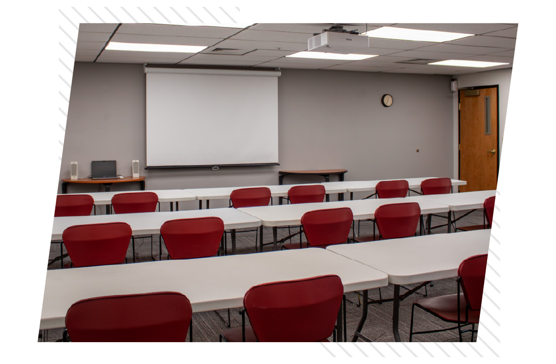 Classroom with rows of white tables and red chairs facing a ceiling-mounted projector and pull-down screen, with a laptop and speakers on a front table and a wall clock visible.