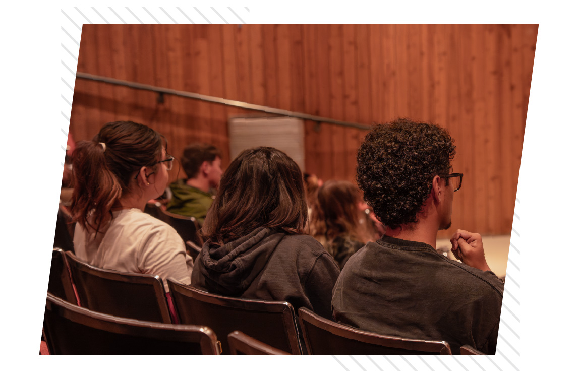 Rear view of several students seated in auditorium chairs, facing forward toward an unseen stage; wood-paneled walls and tiered seating suggest a university lecture or presentation setting.