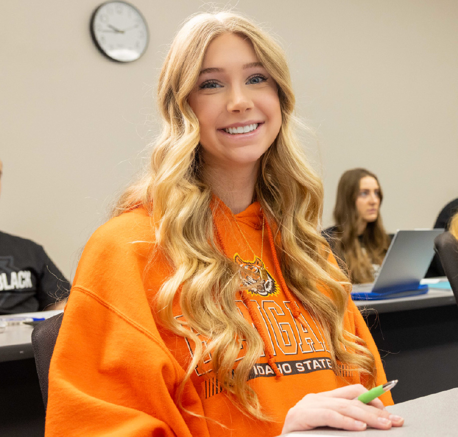 Smiling Idaho State University student wearing an orange Bengals hoodie sits in a classroom holding a pen, with other students and laptops visible in the background.