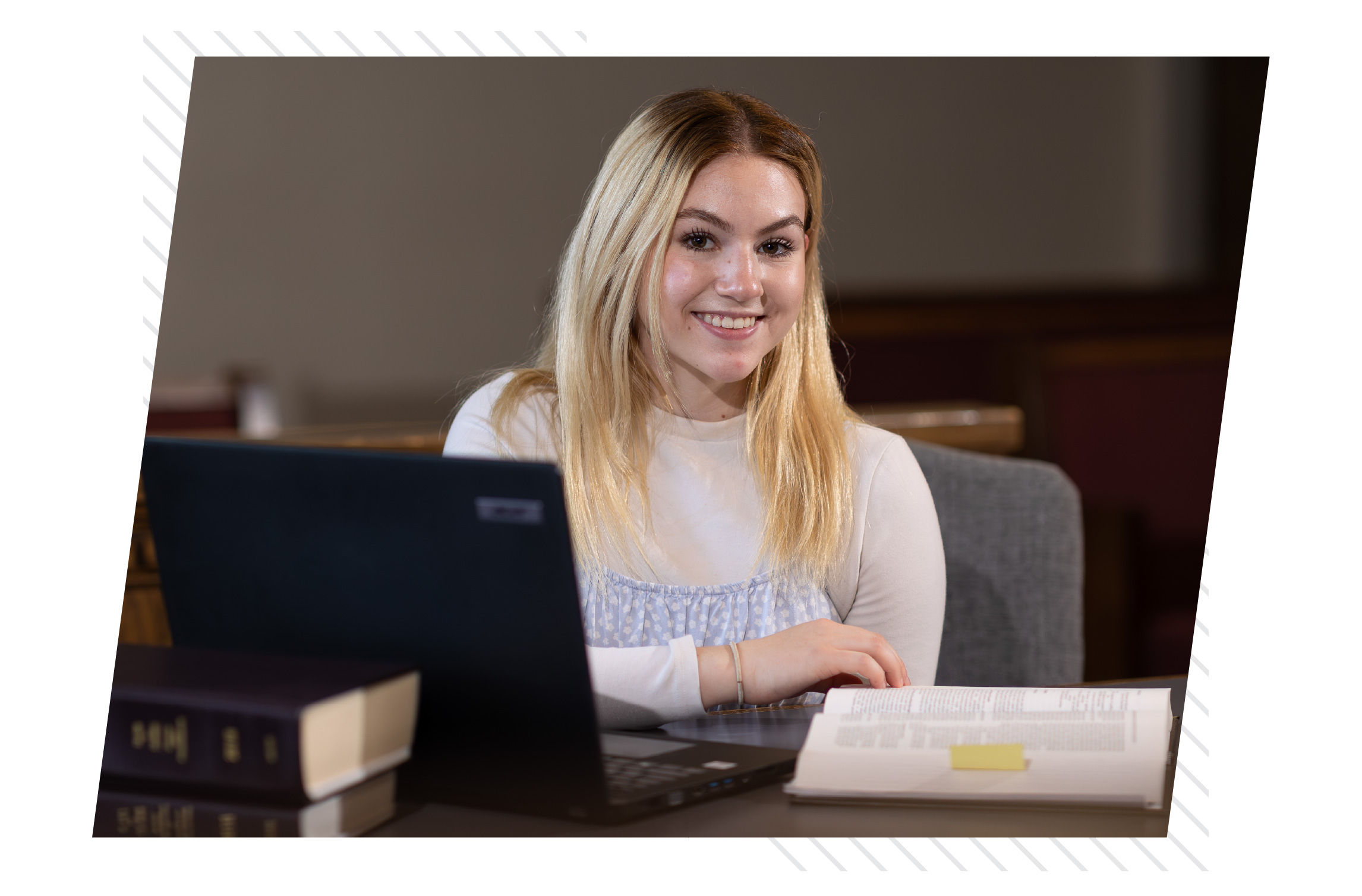 Smiling female college student studying at a table with a laptop and open book in a campus library or study space.