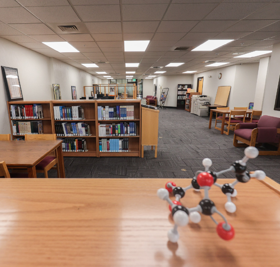 Campus library study area with bookshelves, study tables, and seating, with a molecular model displayed on a table in the foreground.