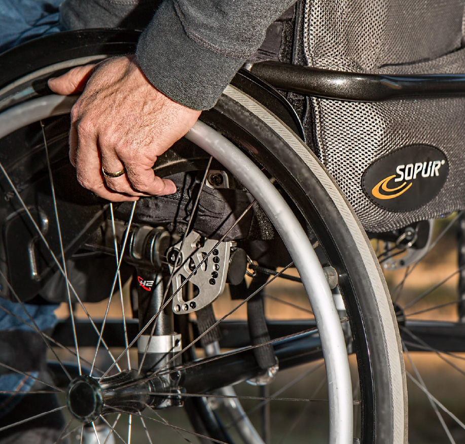 Close-up of a person’s hand resting on the wheel of a manual wheelchair. The image highlights the wheel, spokes, and seating frame, emphasizing mobility and accessibility.