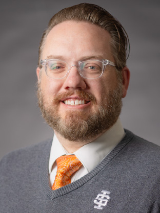 Portrait of a smiling man wearing glasses, a gray sweater with an Idaho State University logo, and an orange patterned tie against a neutral background.