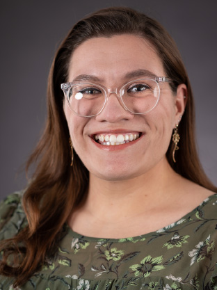 Portrait of a smiling woman wearing glasses and a green floral blouse against a neutral background.