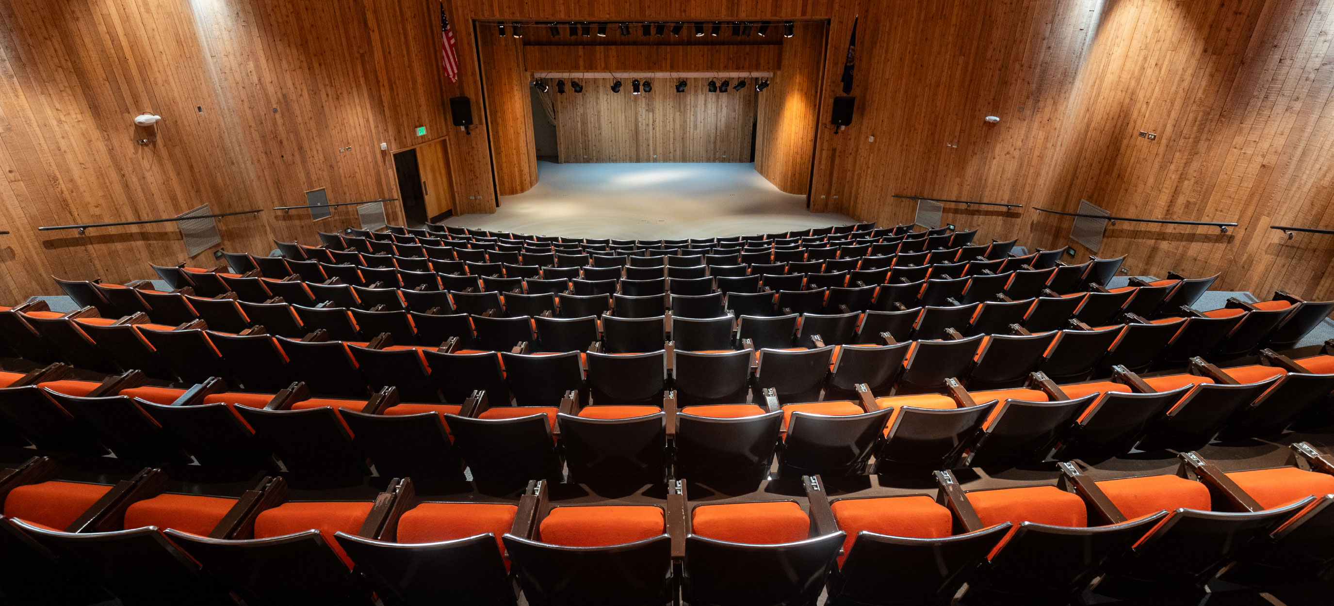 Centered interior view of the TAB Auditorium showing tiered rows of black chairs with orange seats facing a wide, wood-paneled stage with overhead stage lighting; wall-mounted lights and aisle railings frame a professional setting for lectures, performances, and large events.