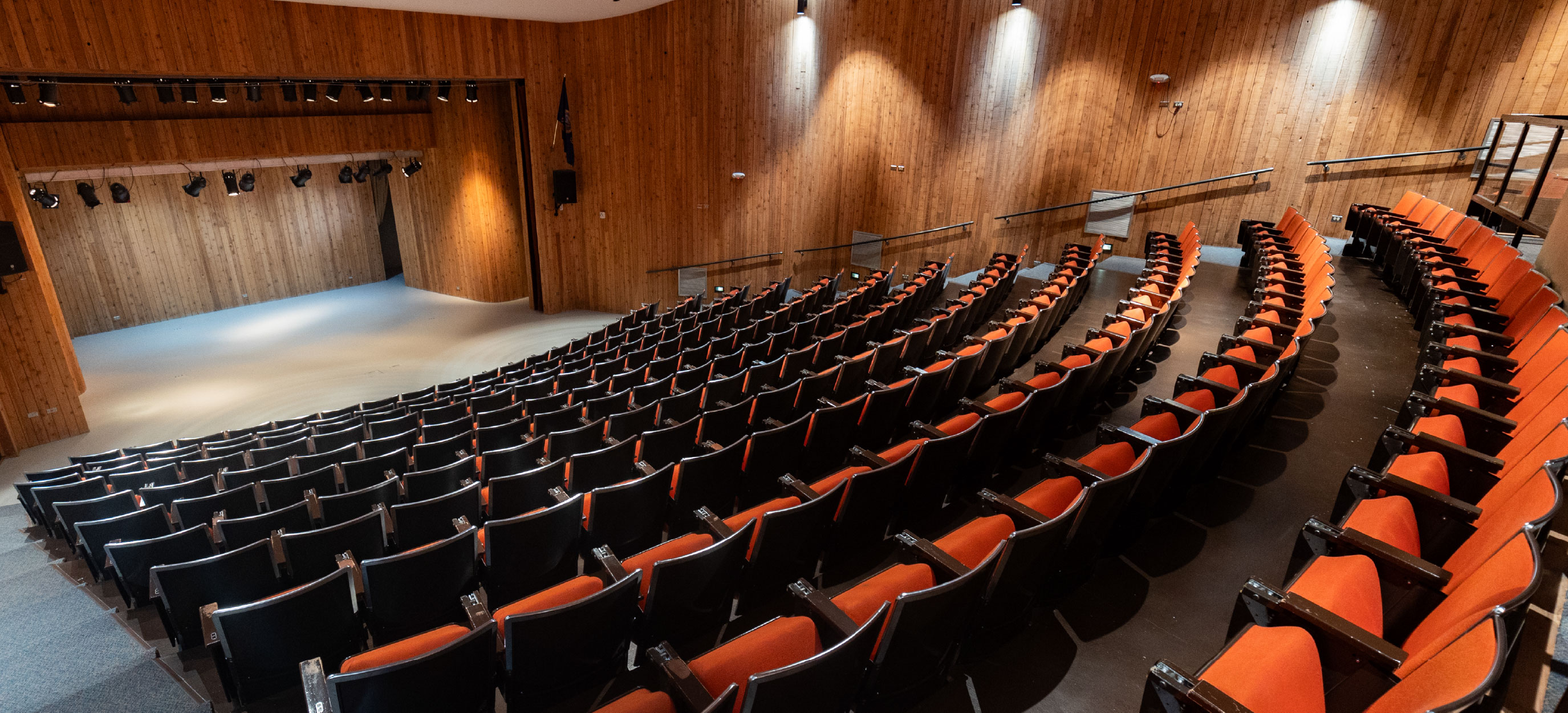 Wide interior view of a tiered auditorium with curved rows of black chairs with orange seats, facing a wood-paneled stage with overhead stage lighting; wall-mounted lights and handrails line the aisles, creating a professional venue for lectures, performances, and presentations.
