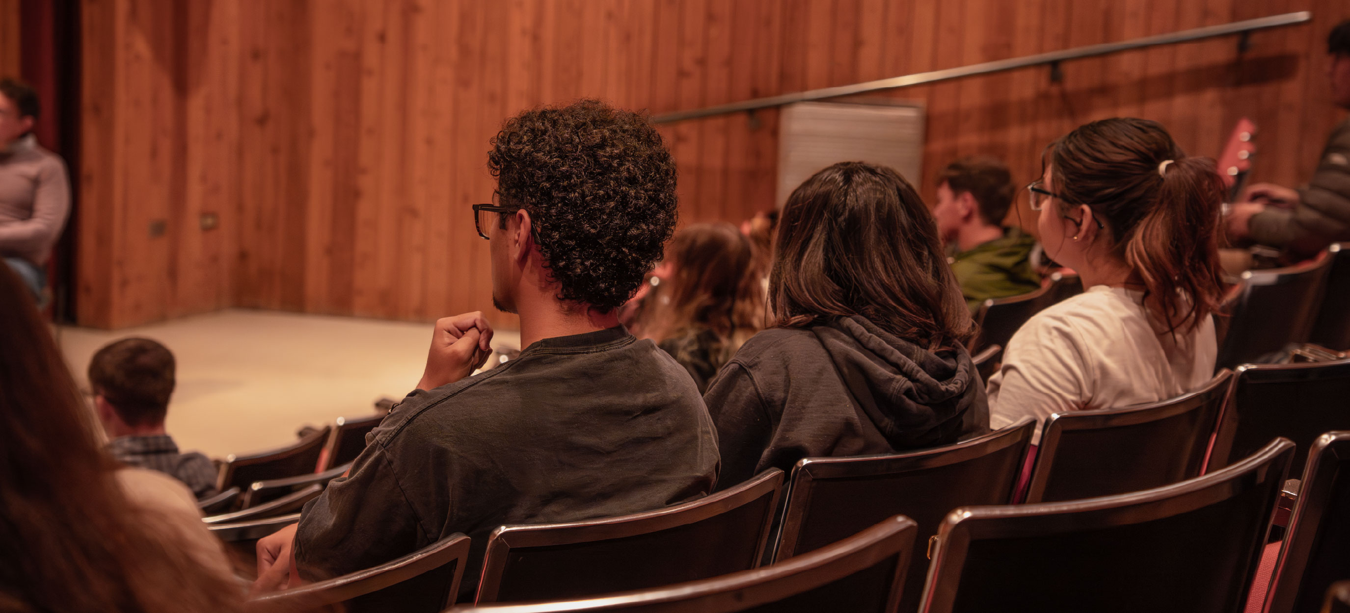 Rear view of several students seated in auditorium chairs, facing forward toward an unseen stage; wood-paneled walls and tiered seating suggest a university lecture or presentation setting.