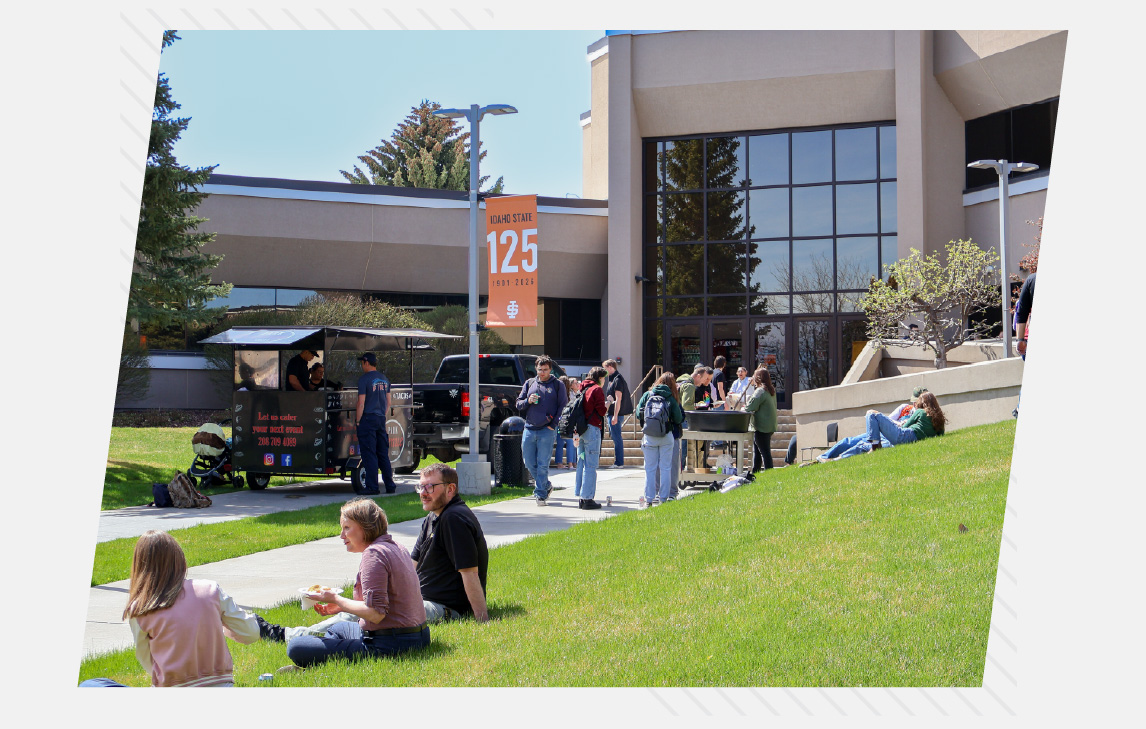 Students gather outside an Idaho State University Idaho Falls campus building on a sunny day, sitting on the grass, walking along the sidewalk, and lining up at a food truck near the entrance.