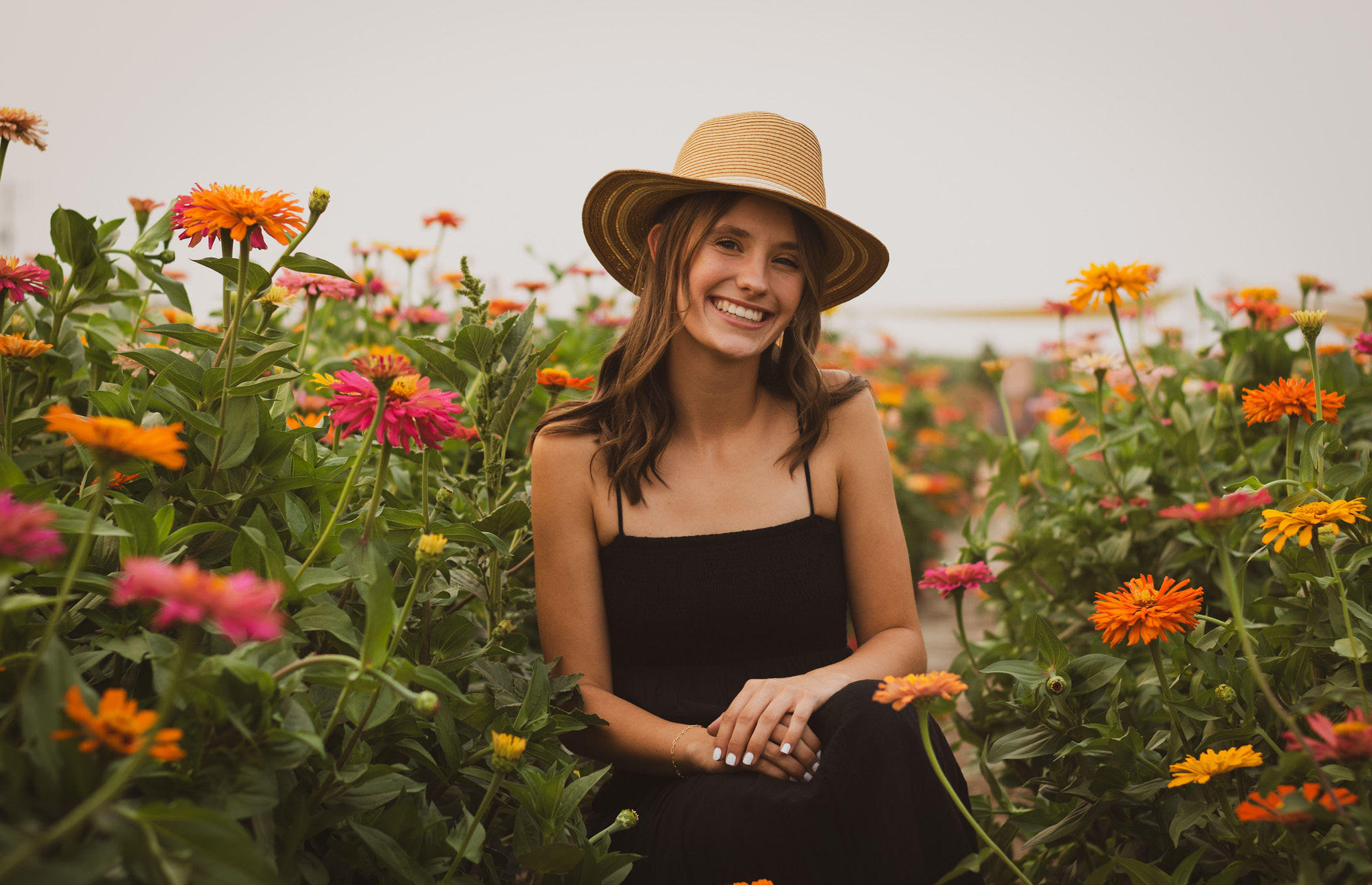 A young woman with a joyful, wide smile sits in a field of colorful zinnias. She is wearing a wide-brimmed straw hat and a black spaghetti-strap dress. She is surrounded by pink, orange, and yellow flowers, with her hands resting gently in her lap. The background is a soft-focus garden under warm, diffused light.