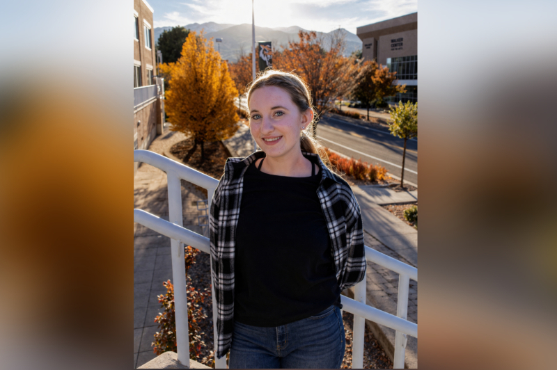 A young woman with blonde hair and blue eyes smiles while leaning against a white metal railing on an elevated walkway. She is wearing a black t-shirt, a black and white plaid flannel shirt, and blue jeans. The background shows a university campus during autumn, with vibrant orange trees, a road, and mountains in the distance under a bright sun that creates a soft glow around her.